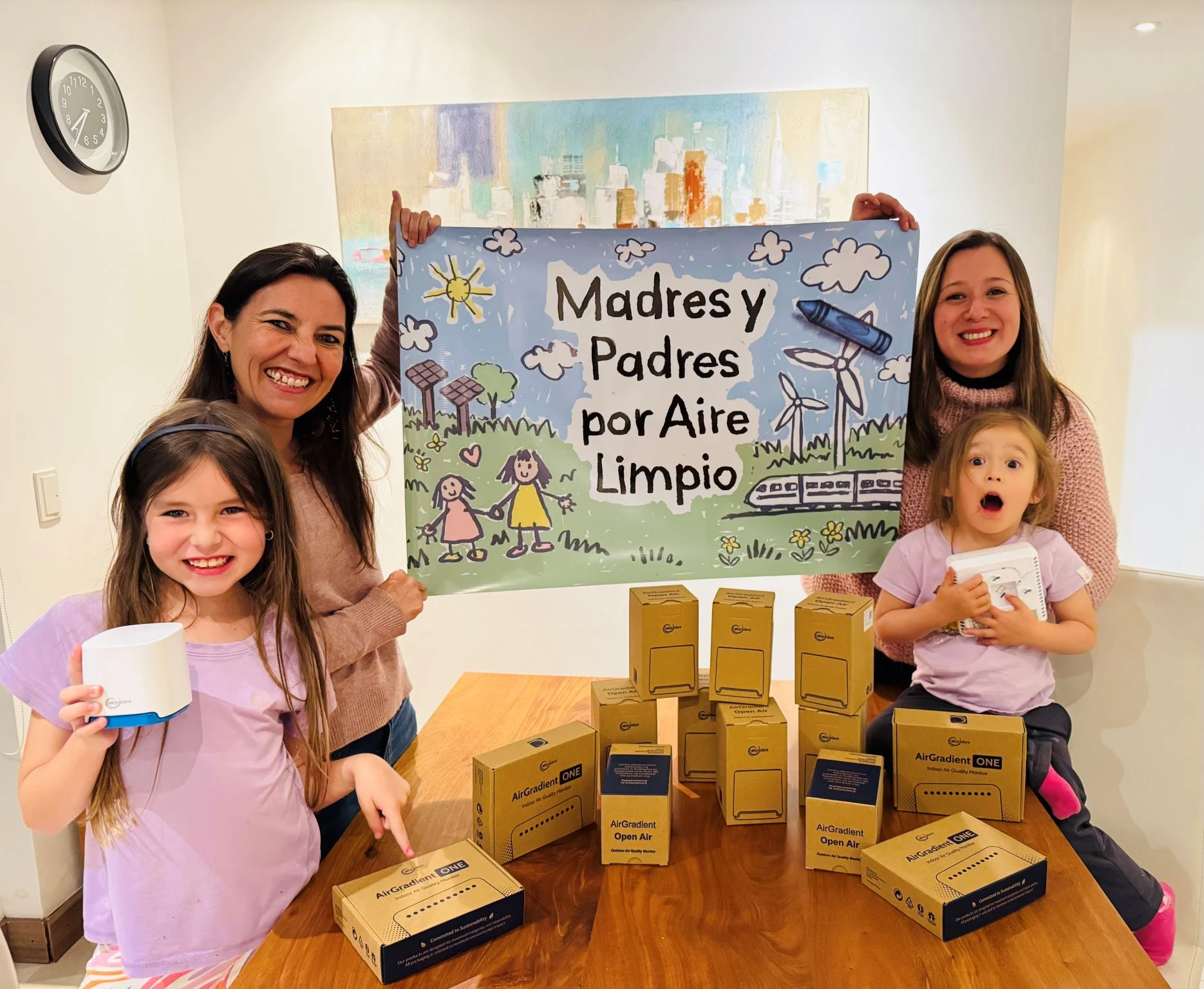 Parents and children holding clean air signs with AirGradient monitor boxes on a table.