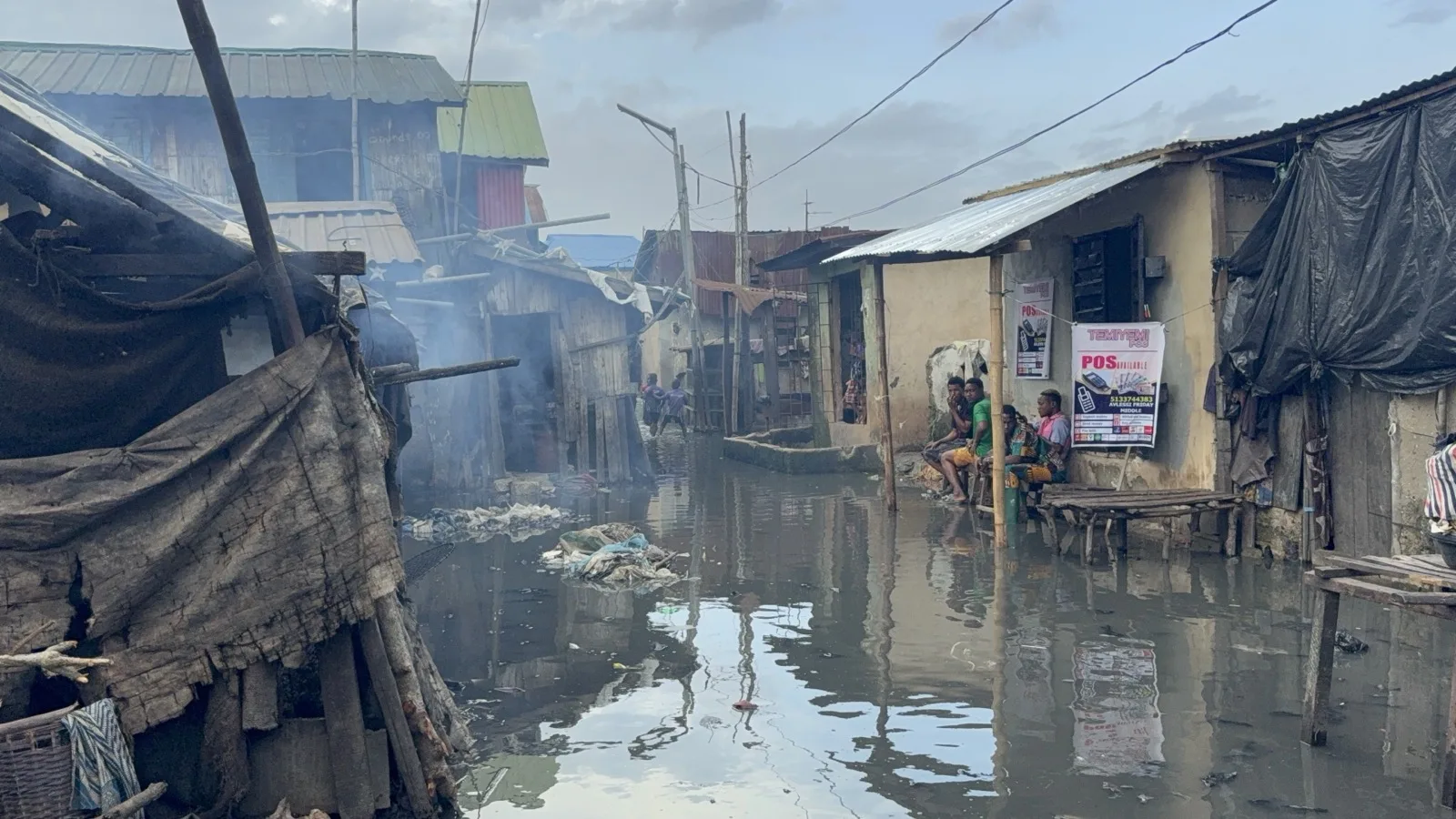 Flooded Lagos community with homes, smoke, and residents near the water.